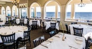 White tablecloth covered dining tables with black chairs and arched windows facing the ocean.